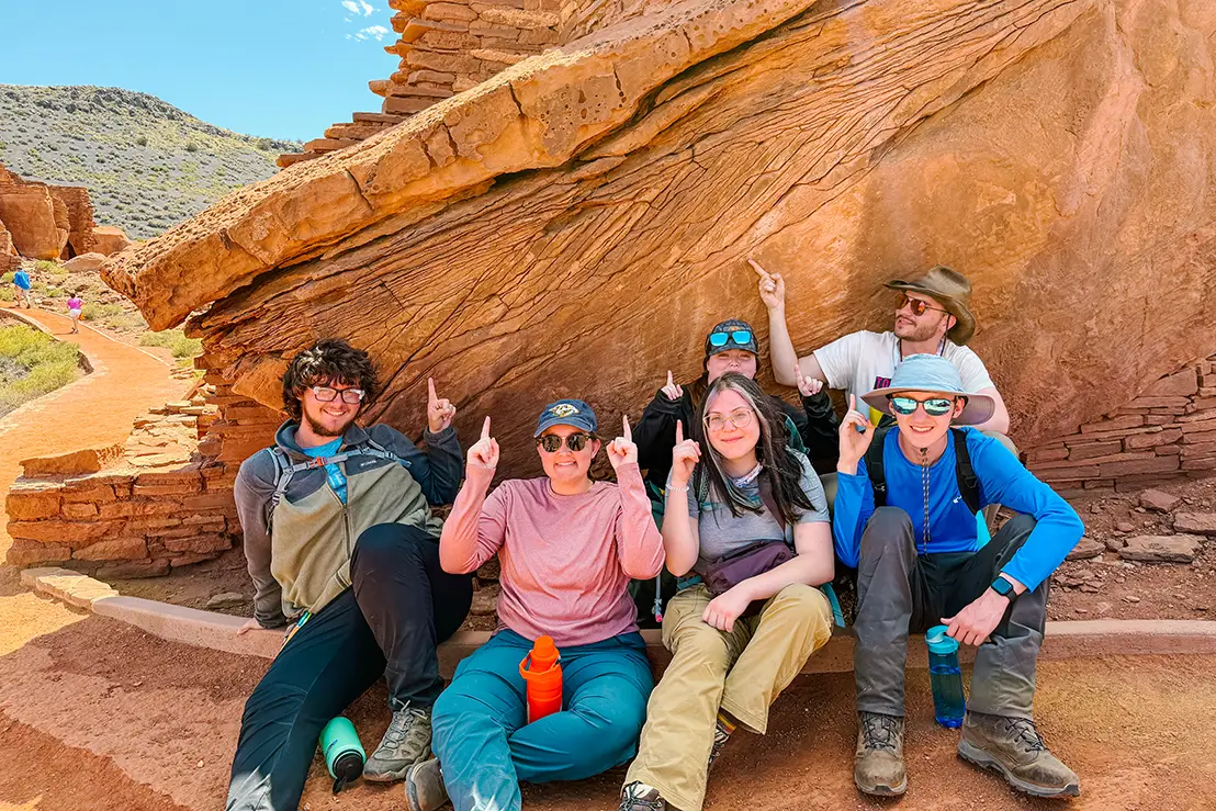 Students from the Department of Earth, Environmental, and Planetary Science point at a rocky red mountain that they are sitting against.