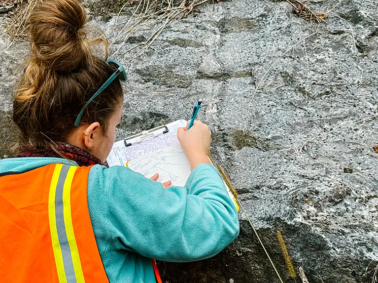 A student draws a geologic pencil drawing while working in the field on mountainous rocks.